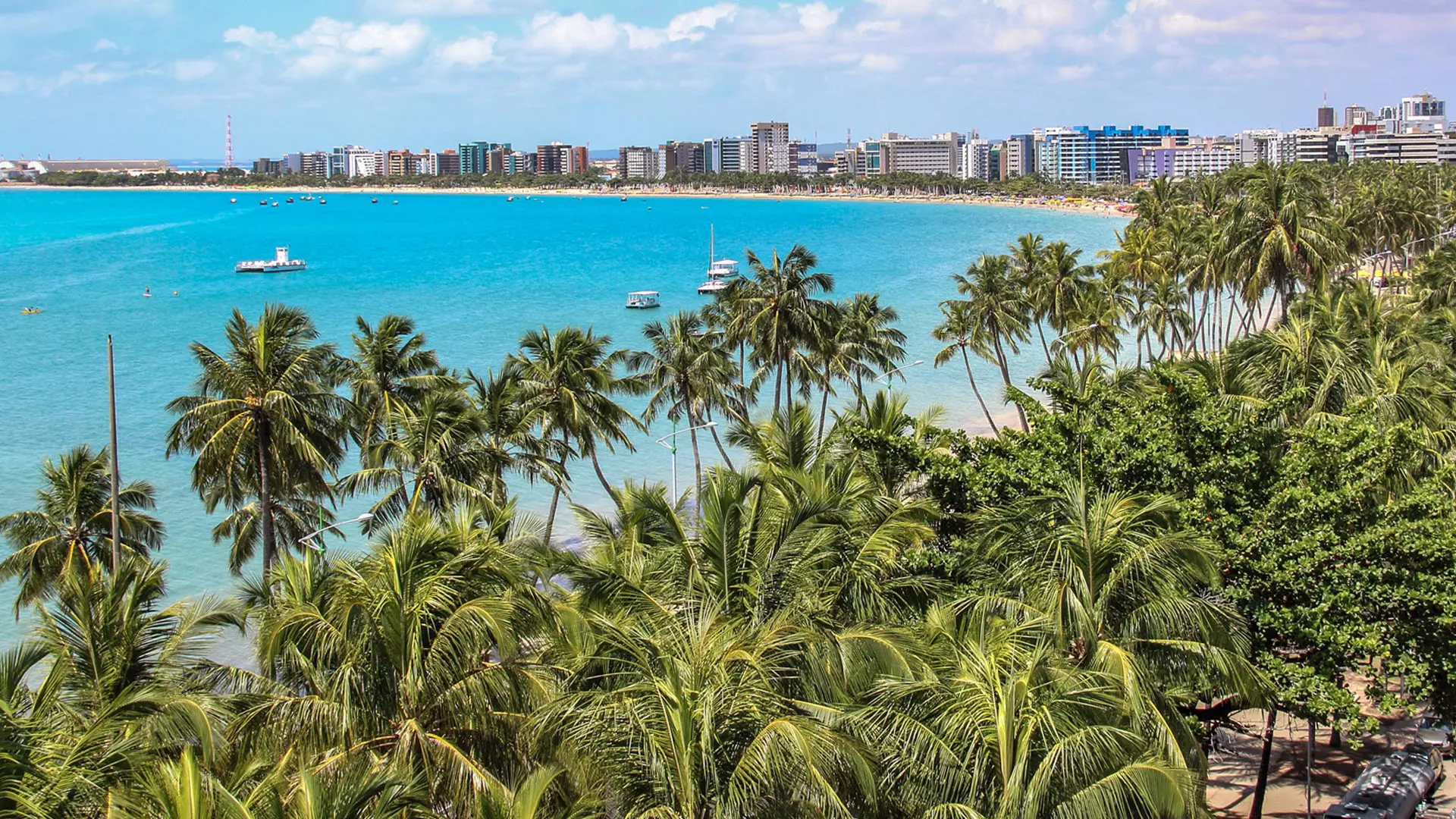 Vista aérea de orla urbana de cidade nordestina com mar azul, palmeiras e prédios ao fundo.
